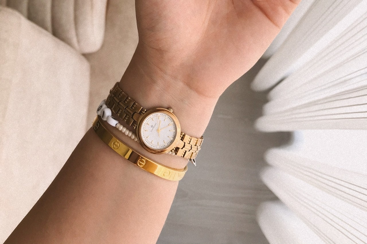 A person’s arm wearing a gold watch, gold bangle, and a white beaded bracelet