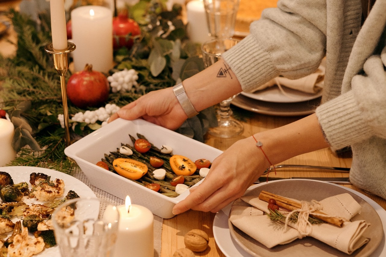 A woman wearing a cardigan and bracelets sets a cassrole dish on the Thanksgiving table.