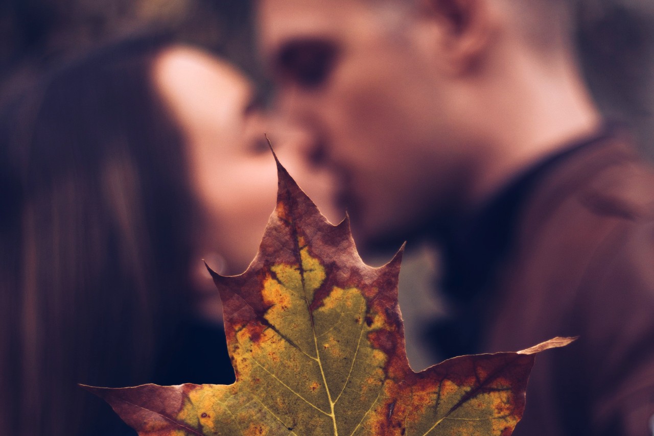 A newly-engaged couple kissing out of focus, with a crisp fall leaf in the foreground.