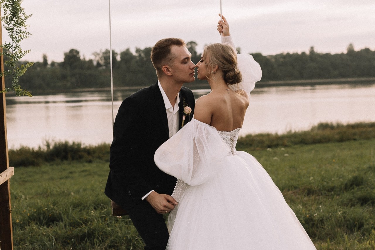 A bride and groom kissing on a wooden swing in front of a calm lake.