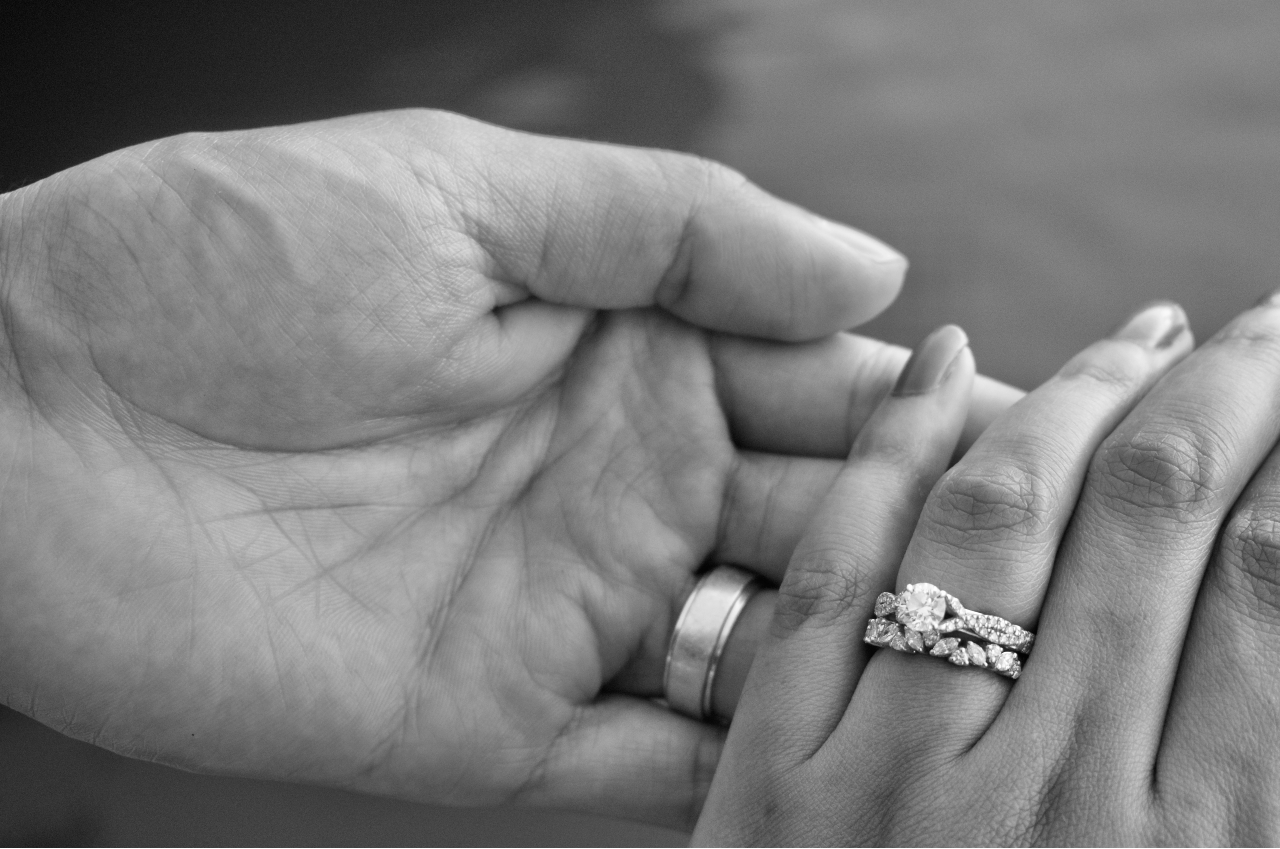 a lady wearing a round-cut engagement ring and wedding band holding a man’s hand